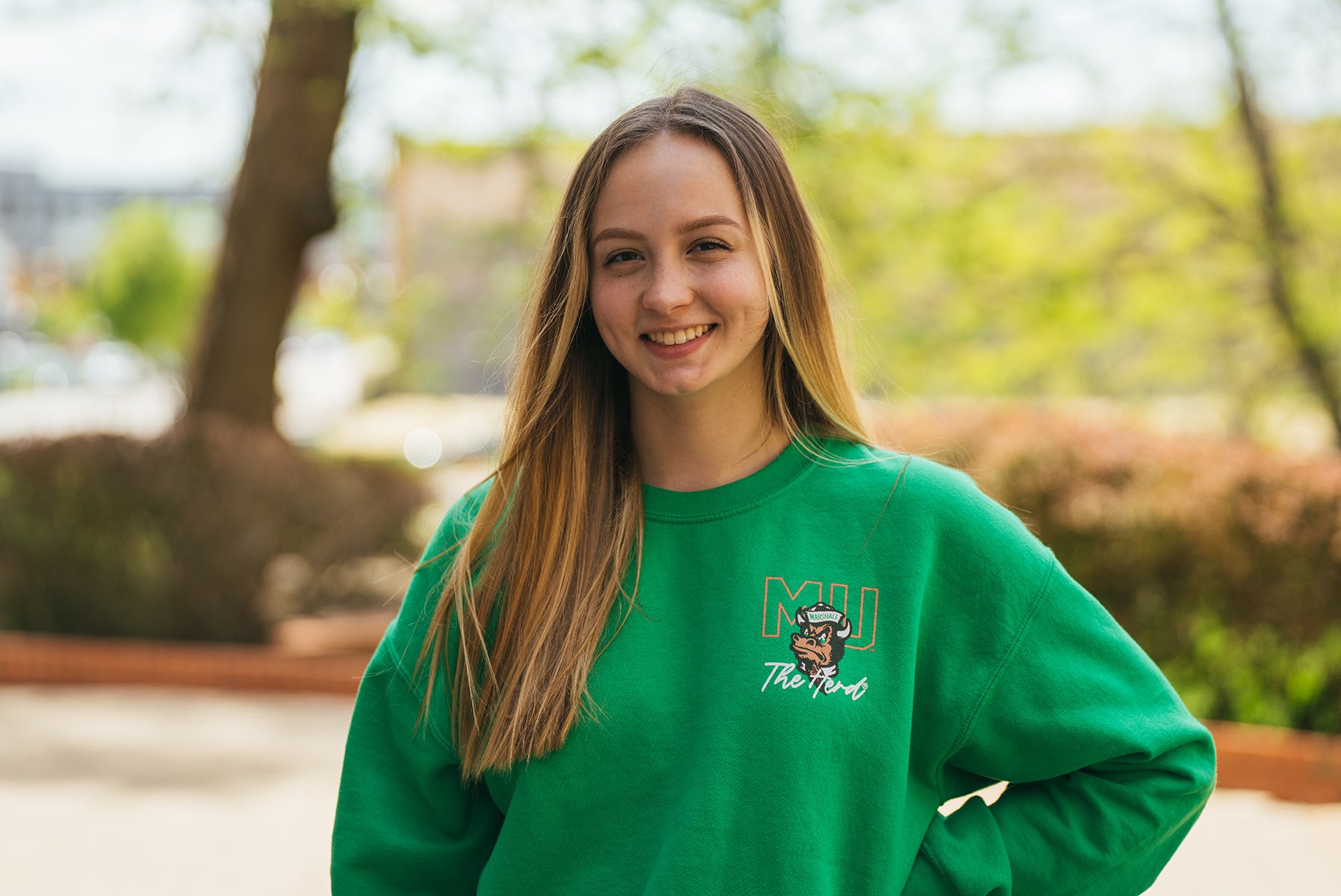 Liv Stockwin wears a green, Marshall sweatshirt as she smiles at the camera from Marshall University's campus
