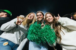 Three Marshall University students pose with green pom-poms and make the Herd hand sign at a nighttime football game.