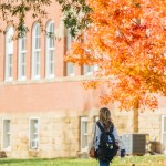 A Marshall University student walks across campus beneath vibrant orange autumn leaves.