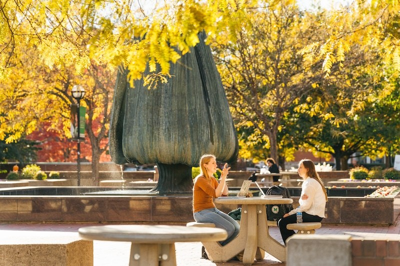 Two Marshall University students sit at an outdoor table near the Memorial Student Center fountain surrounded by colorful fall foliage.