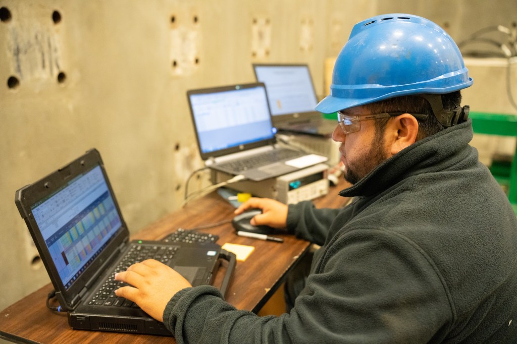 engineering student working on two computers in the materials testing laboratory while wearing a hard hat and safety glasses