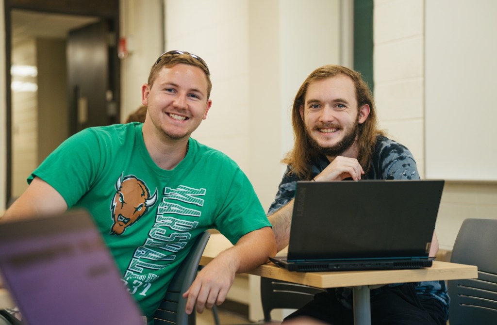 two students in a classroom smiling at the camera