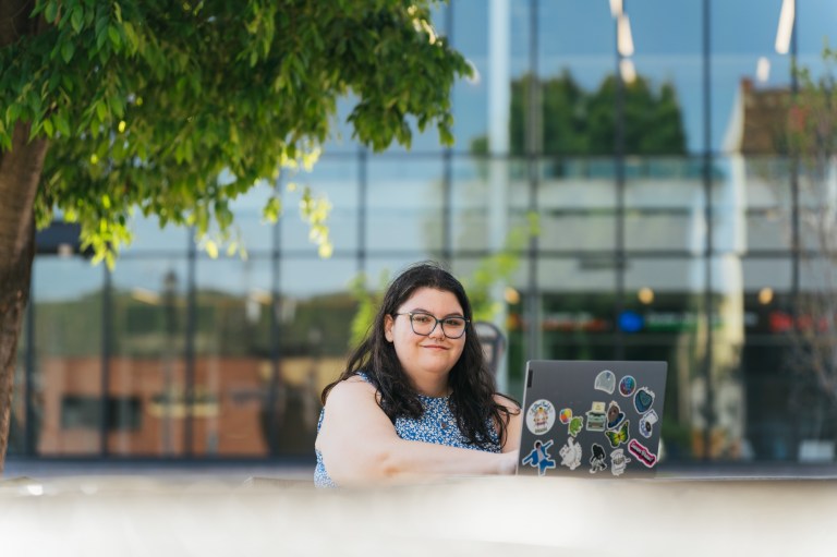 student working on a laptop outside the School of Business Building and smiling at the camera