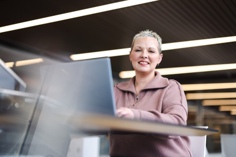 student working at a laptop at a desk and smiling at the camera