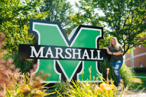Skyla Hilliard with large Marshall Sign on Marshall University's campus.