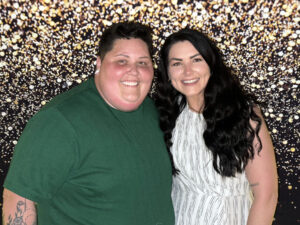 Mika Sheppard and Caylin France smile together for a photo in front of a silver, glitter background
