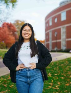 Gracie Queen smiles while wearing a Marshall shirt and standing in front of Marshall University's Drinko Library 