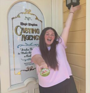 Kelci Armstead excitedly holds out her celebration pin in front of a Magic Kingdom Casting Agency sign.