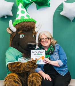 Marco wears a birthday hat while sitting with Author Michelle D. Callanta who is holding her book, Let's Cheer with Marco. Balloons are behind them.