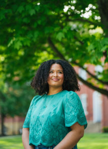 Haleigh Muncy wears a green shirt and stands under a tree looking up with a red brick building in the background