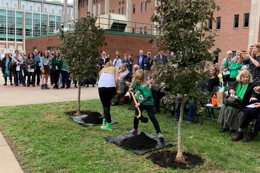 Participants take part in a memorial tree planting ceremony between the Science Building and Harris Hall, placing soil around newly planted trees while attendees gather nearby.