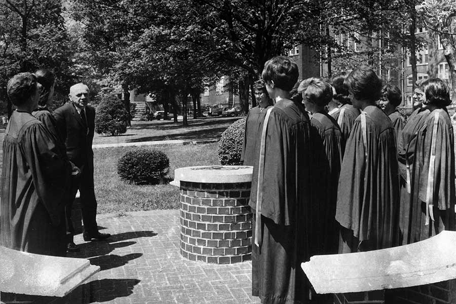 Historic black-and-white photograph of an Omicron Delta Kappa induction ceremony at the ODK Circle near Old Main. A group of robed initiates stands within the circular brick gathering space while a university official addresses the ceremony.