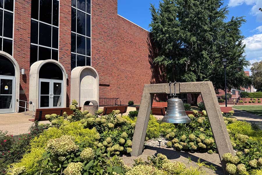 Thornburg Bell displayed in a landscaped plaza south of Morrow Library, with the commemorative bronze bell suspended from a concrete frame and surrounded by benches, flowering plantings, and campus walkways.