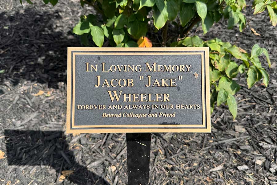 Close-up of a memorial plaque beside a flowering dogwood tree reading, “In Loving Memory Jacob ‘Jake’ Wheeler, Forever and Always in Our Hearts, Beloved Colleague and Friend.”