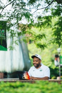 Marshall University staff member works on a laptop outdoors beneath leafy trees on a sunny day on campus.