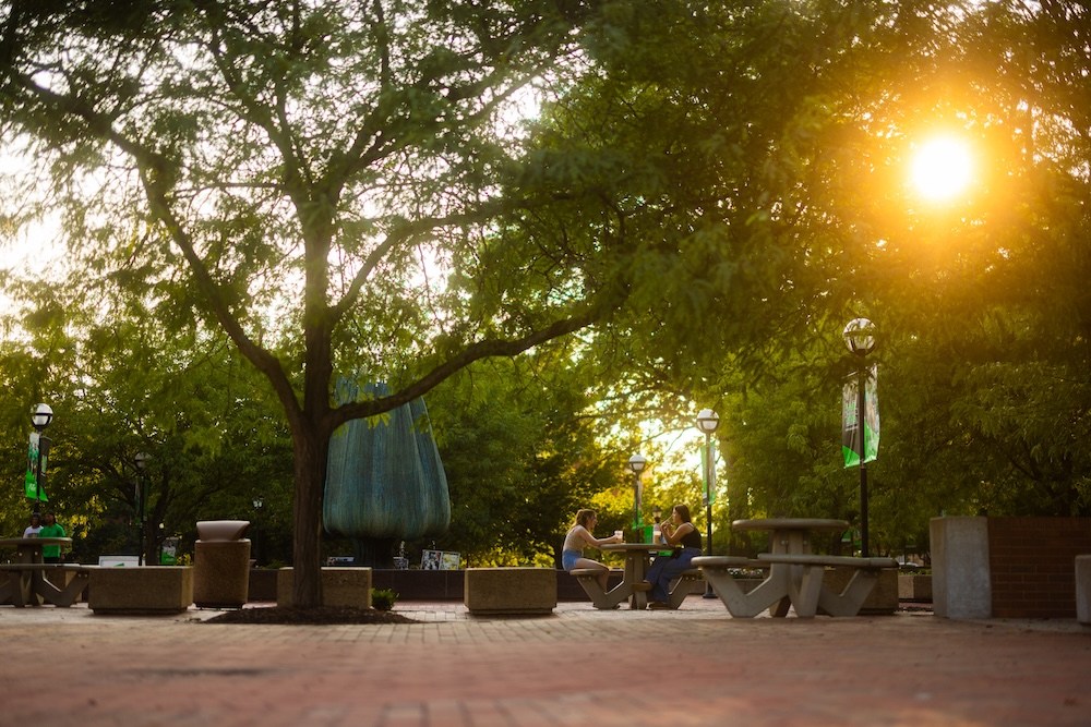 Students sit and talk at outdoor tables in a campus plaza during sunset at Marshall University.
