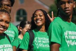 Smiling students wearing green “Experience My Marshall Moment” shirts walk together during a campus event.